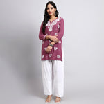 Woman wearing a purple embroidered top and white pants on a gray background