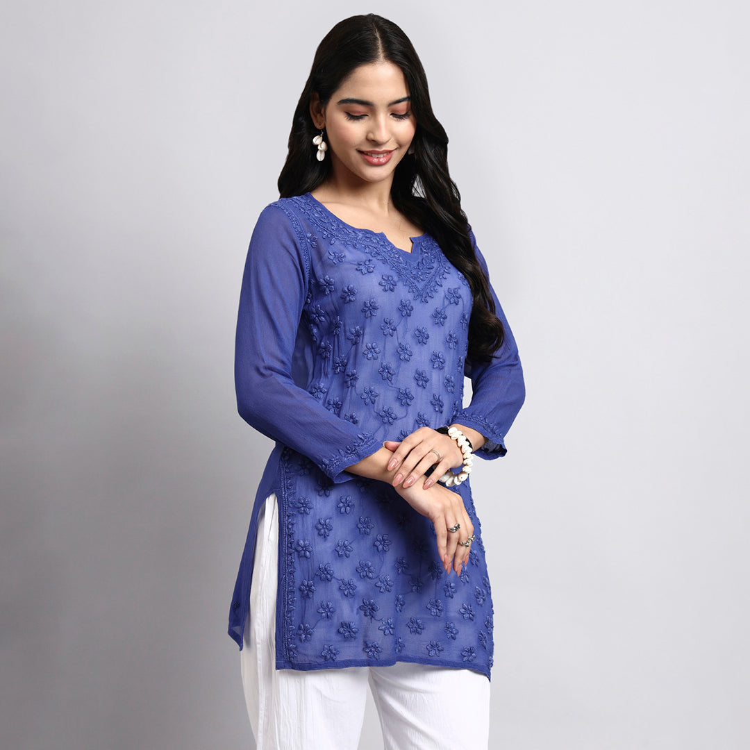 Woman wearing a blue embroidered top with white pants on a gray background