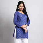 Woman wearing a blue embroidered top with white pants on a gray background