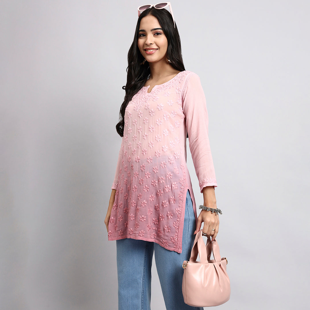 Woman wearing a pink short kurta with floral patterns and blue jeans, holding a pink handbag against a gray background.