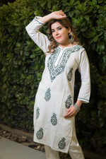 In a front view, a woman with her hand raised wears a white kurti with stylized green motif prints.