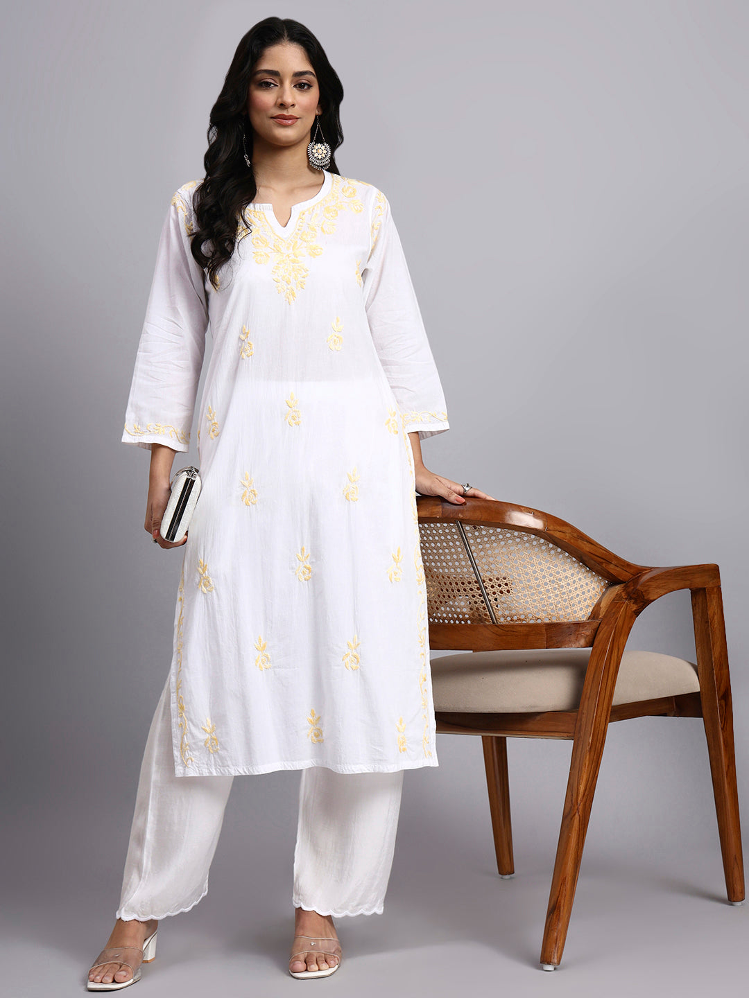 Full view of Woman wearing a white kurta with yellow patterns standing next to a wooden chair on a gray background
