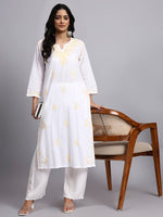 Full view of Woman wearing a white kurta with yellow patterns standing next to a wooden chair on a gray background