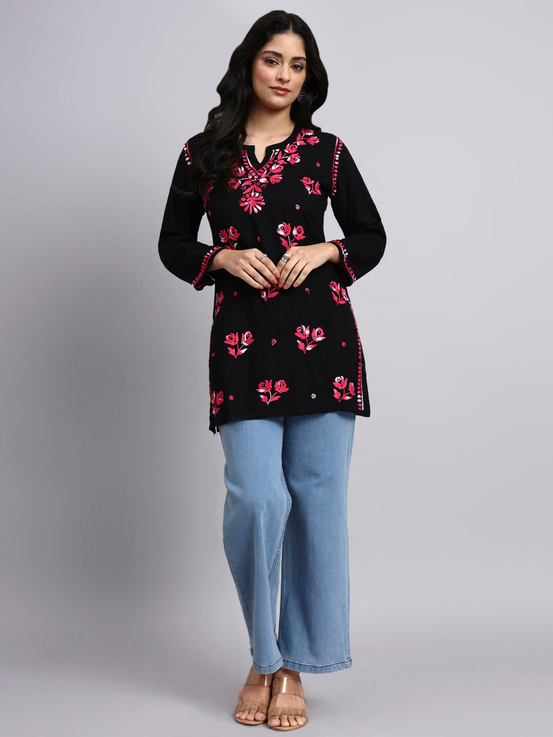 Full view of Woman wearing a black floral embroidered top and light blue pants on a gray background