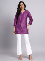 Woman wearing a purple embroidered short chikankari kurti and white pants on a gray background