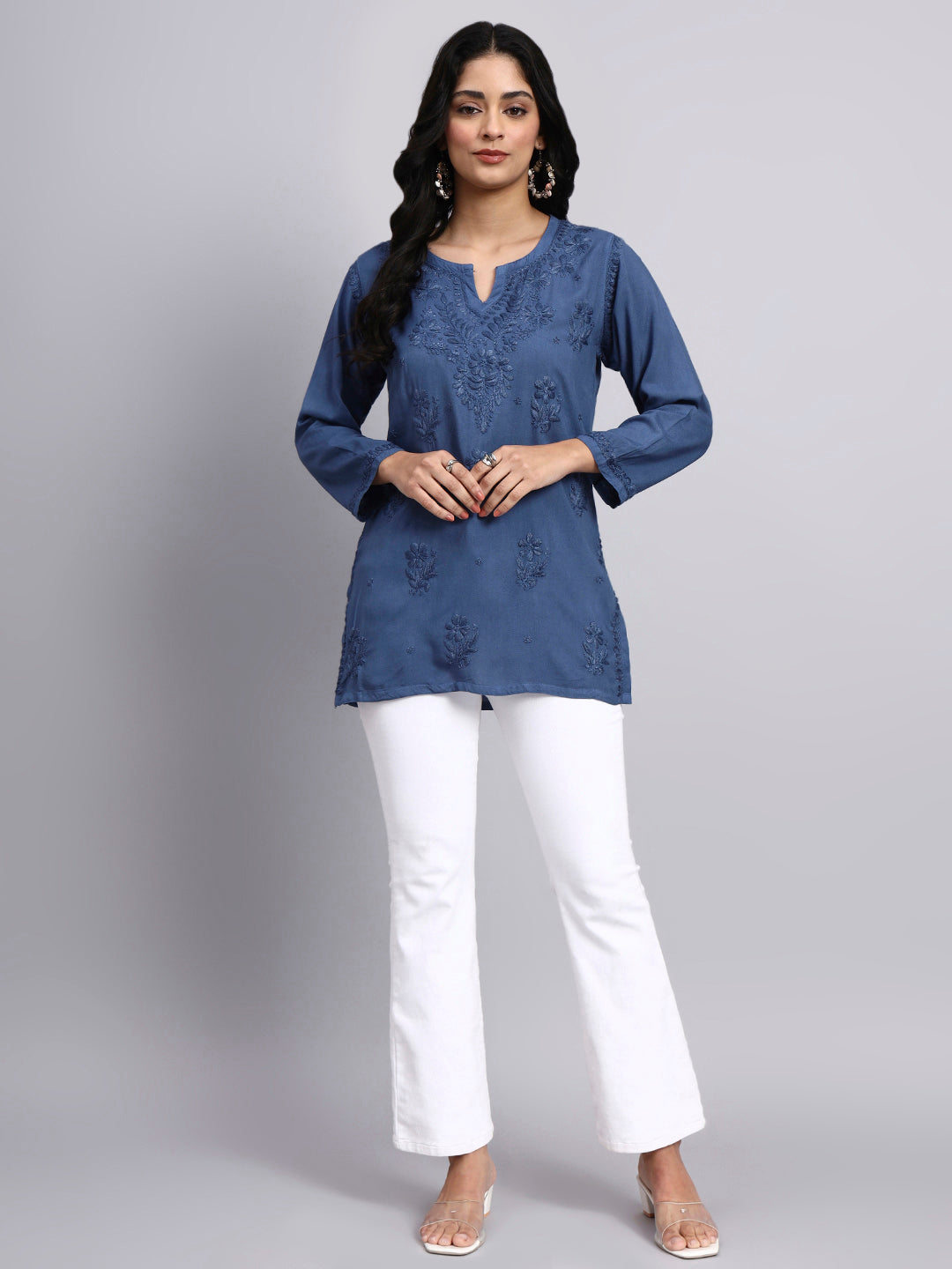 Full look of Woman wearing a blue embroidered top and white pants on a gray background