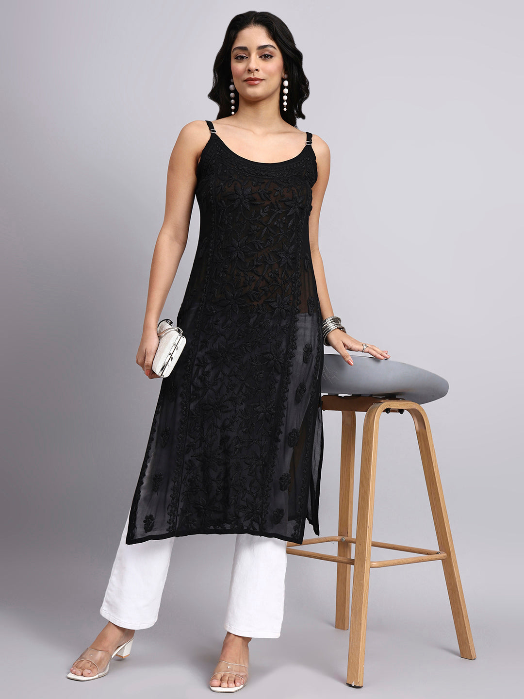 Woman wearing a black sleeveless chikankari kurti and white pants, sitting on a gray stool against a gray background.