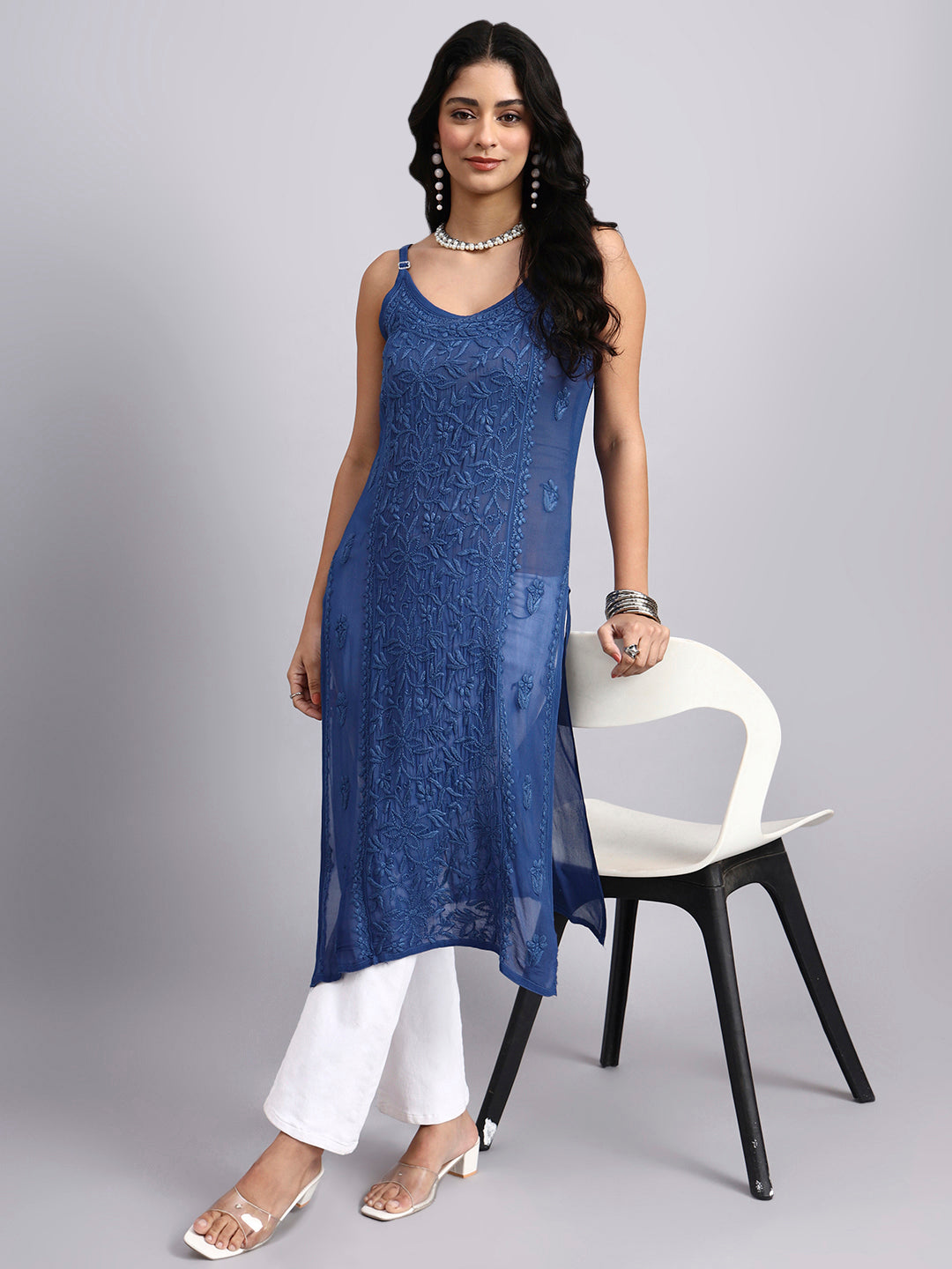 Front look of Woman wearing a blue sleeveless dress with white pants, standing next to a chair on a gray background.