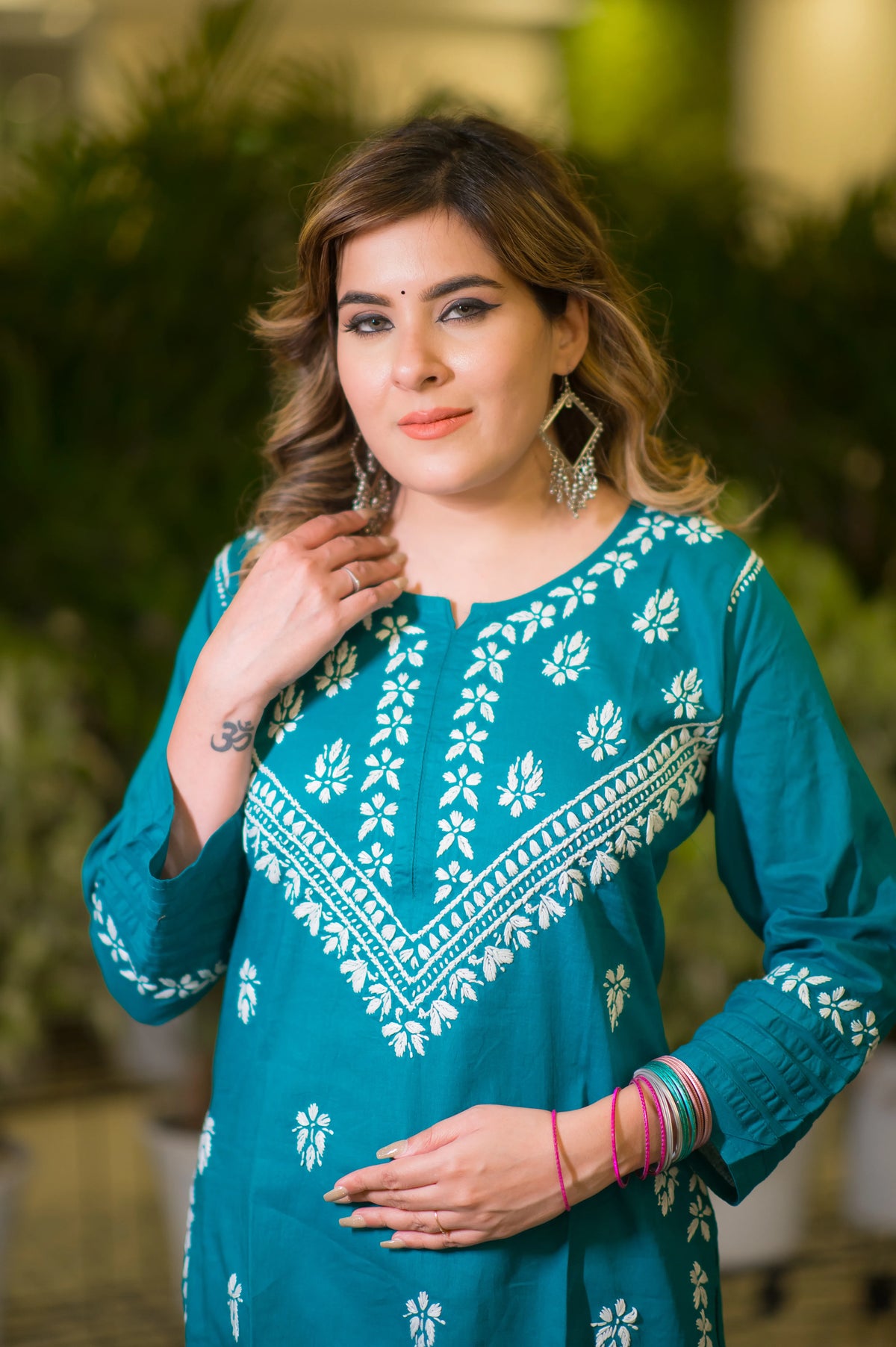 In a close-up front view, a woman touching her hair wears a teal blue kurta with white floral and geometric embroidery. 