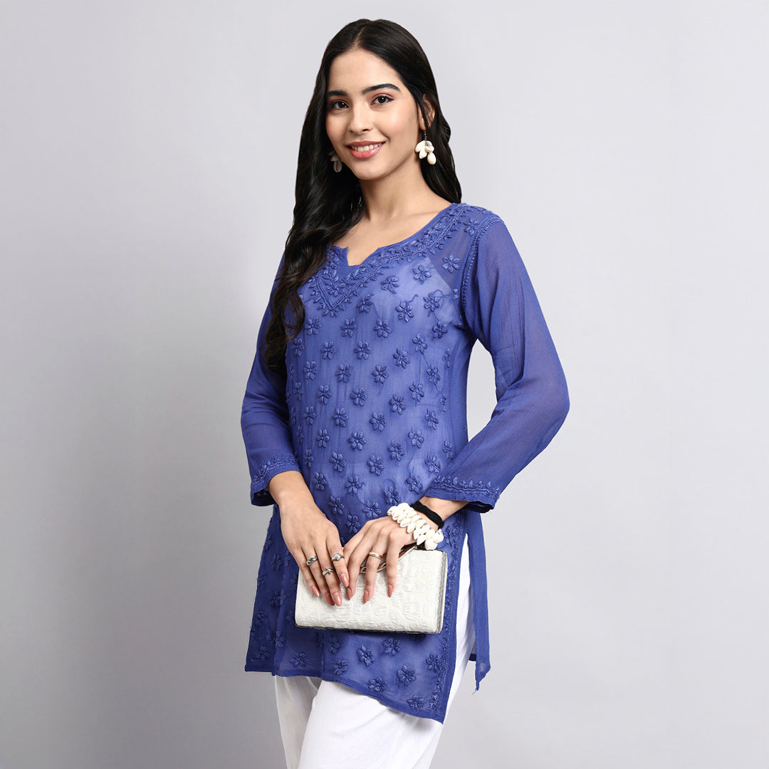 Woman wearing a blue embroidered top and white pants against a gray background