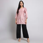 Woman wearing a pink embroidered top and black pants on a plain background