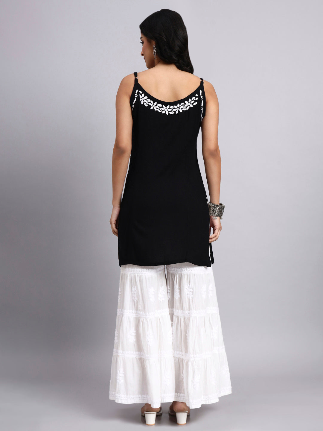 Back view of Woman wearing a black sleeveless top with white embroidery and white checkered pants on a gray background