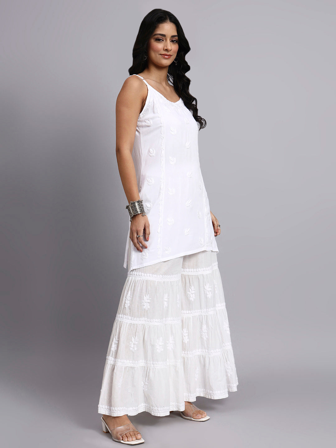 Woman wearing a white sleeveless chikankari kurti set and layered white sharara against a gray background