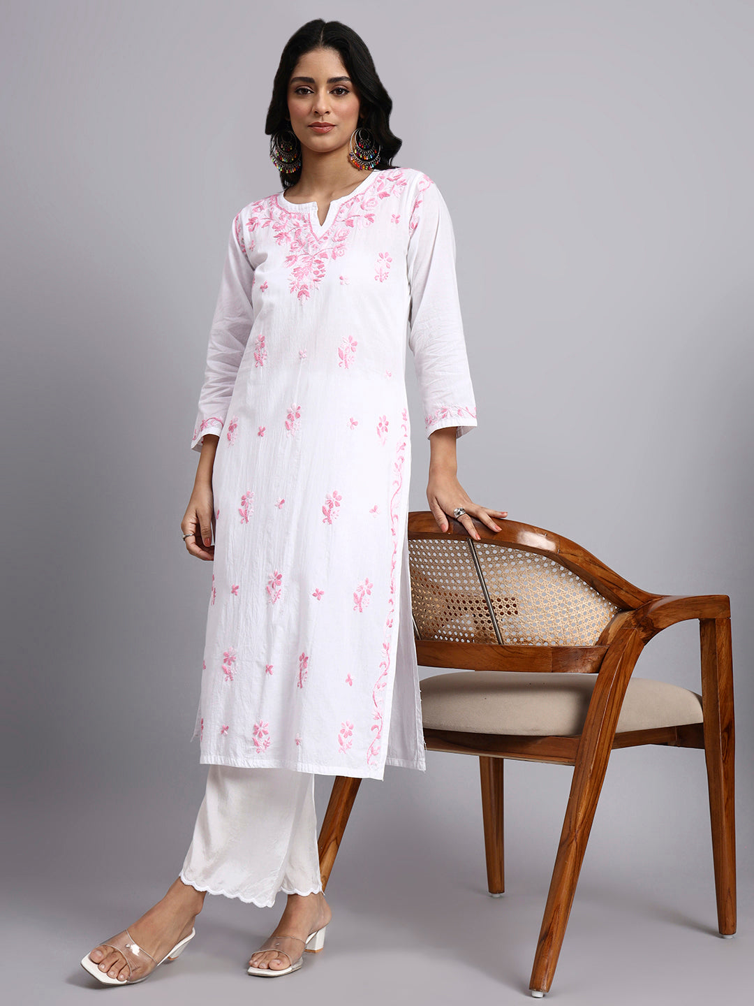 Full front look of Woman wearing a white kurta with pink floral patterns, standing next to a wooden chair on a gray background.