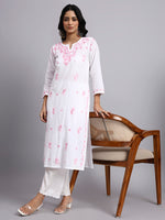 Full front look of Woman wearing a white kurta with pink floral patterns, standing next to a wooden chair on a gray background.