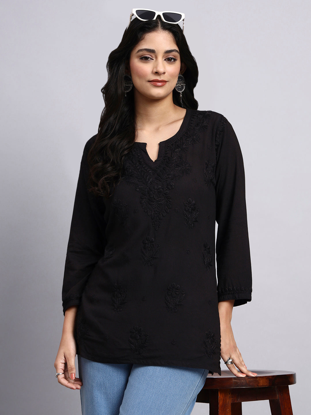 Woman wearing a black embroidered top with blue jeans on a gray background