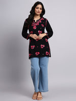 Full view of Woman wearing a black floral embroidered top and light blue pants on a gray background