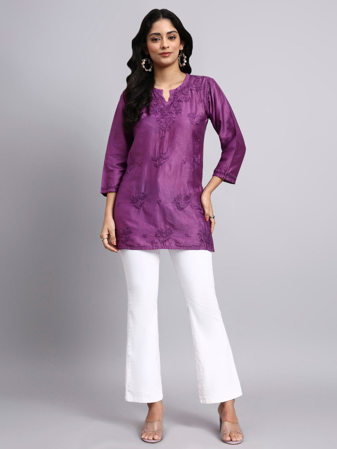 Woman wearing a purple embroidered short chikankari kurti and white pants on a gray background