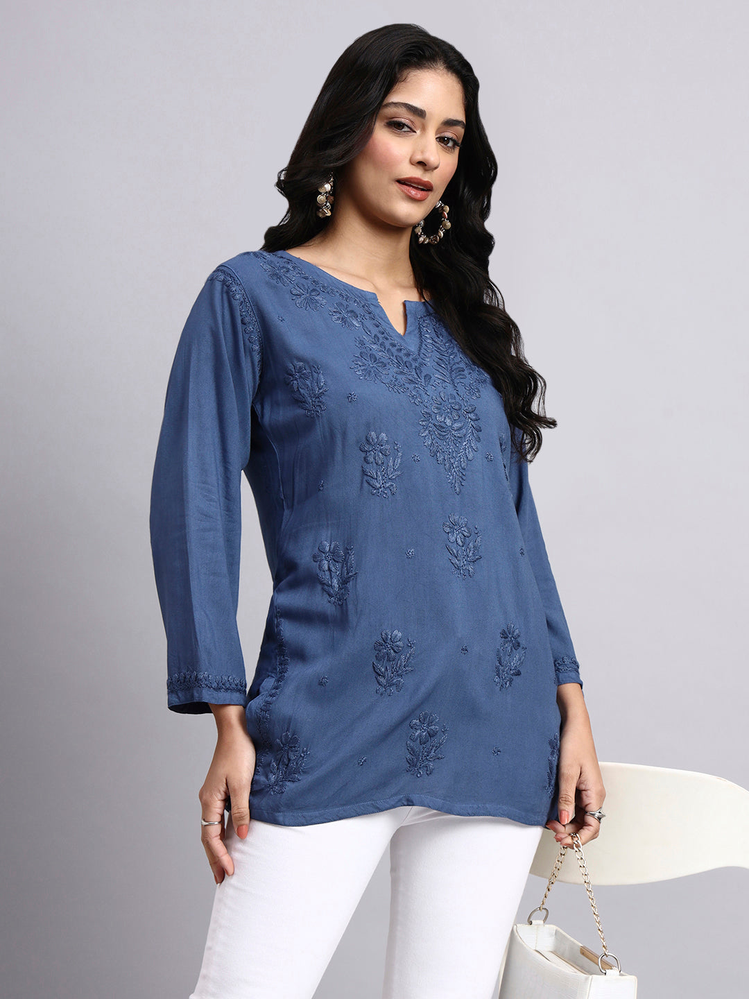 Woman wearing a blue embroidered top with white pants on a gray background