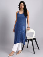 Front look of Woman wearing a blue sleeveless dress with white pants, standing next to a chair on a gray background.
