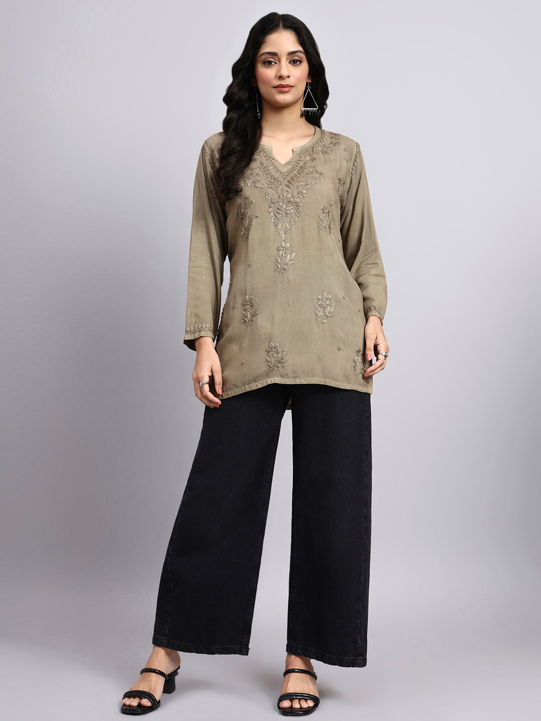 Full view of Woman wearing a olive embroidered top and black pants on a gray background