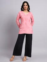 Full view of Woman wearing a pink embroidered top and black pants on a gray background.