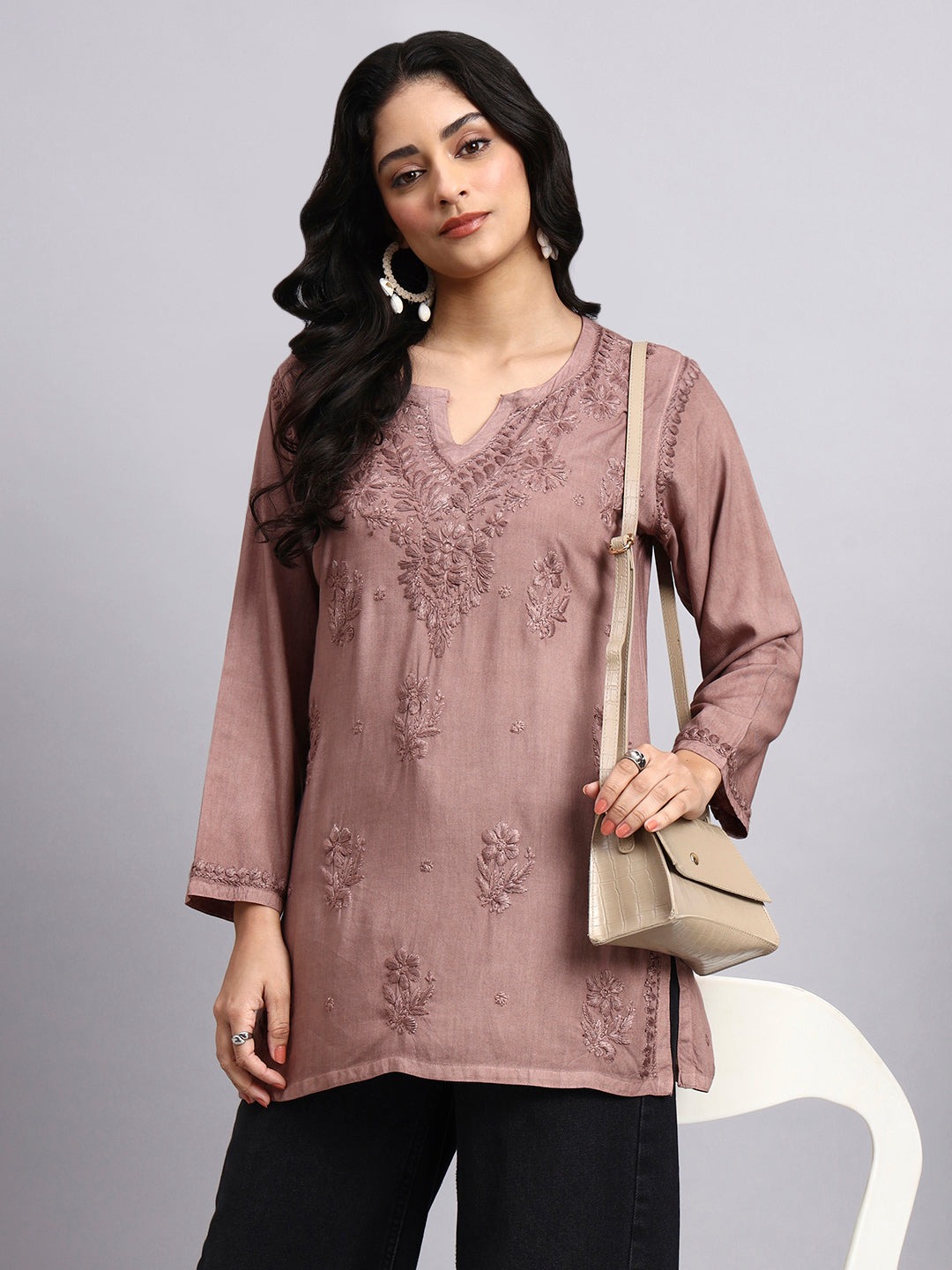 Woman wearing a mauve embroidered short kurta holding a beige handbag against a gray background