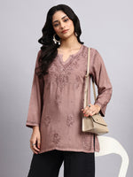Woman wearing a mauve embroidered short kurta holding a beige handbag against a gray background